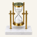 A decorative brass hourglass with a white marble base, featuring a clear glass hourglass filled with white sand and a green top cap.
