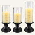 Three black candle holders with graduated clear glass cylinders, each holding a cream-colored pillar candle with a glowing flame and wrapped with thin wire string lights, shown on a white background.