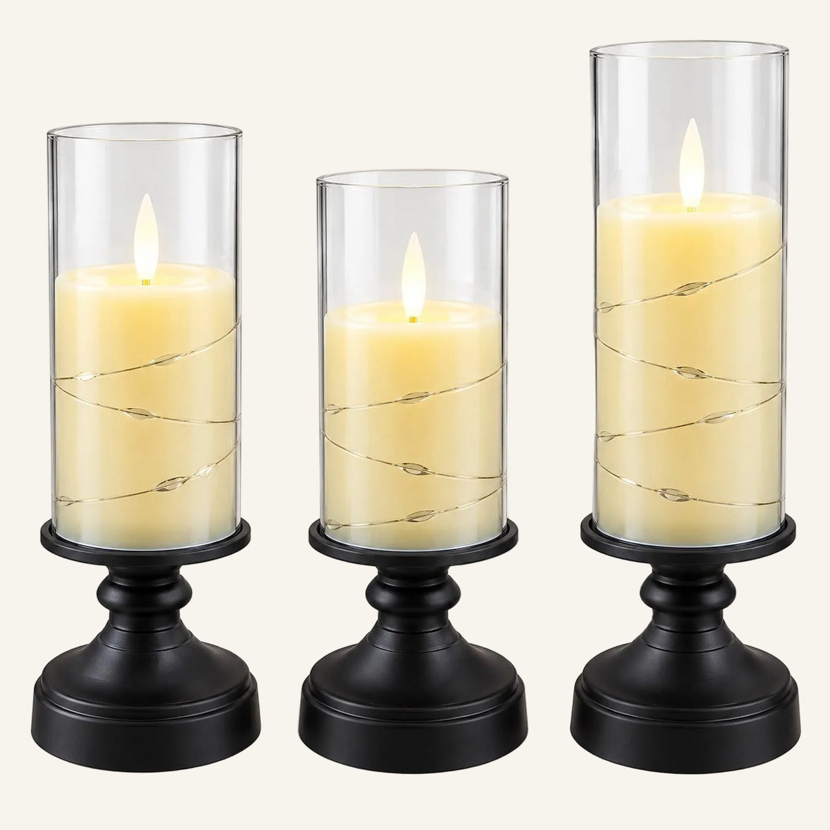 Three black candle holders with graduated clear glass cylinders, each holding a cream-colored pillar candle with a glowing flame and wrapped with thin wire string lights, shown on a white background.