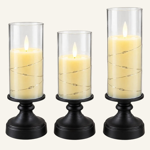 Three black candle holders with graduated clear glass cylinders, each holding a cream-colored pillar candle with a glowing flame and wrapped with thin wire string lights, shown on a white background.