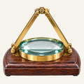 A magnifying glass with a polished brass tripod handle and a dark reddish-brown wooden base, shown on a clean white background.